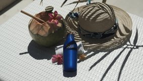 Video footage of green young coconut close up with bamboo straw, bottle of sunscreen SPF 50, sun hat, tropical pink flowers frangipani, palm shade on white sun bed in Bali 