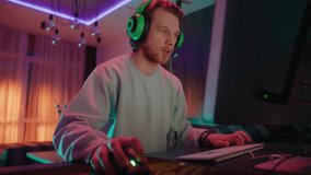 Emotional young attractive man with headphones play games on computer at home in the evening. Videogame, cyber, entertainment, player, technology, internet, streaming, hobby. Slow motion