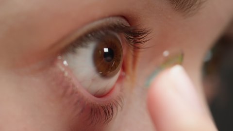 Close-up of a brown-eyed young woman putting on the decorative lens with fingers. Optical shop concept. Advertising of contact colorful lenses for make-up and performances, changing color of eyes.