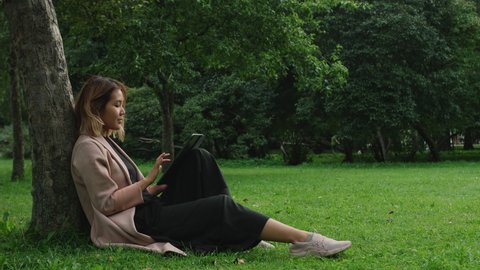 Young asian woman sitting on lawn in city park and working using laptop or tablet computer. Female freelancer works outdoors leaning on a tree on summer day