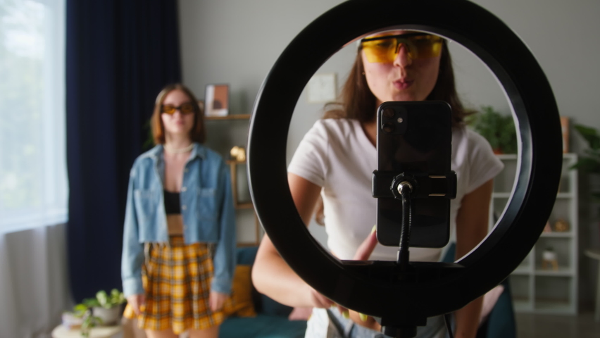 Two young sisters wearing sunglasses recording trend dance video on smartphone with lamp in living room. Female students, teenagers dancing for musical clip. Bloggers making music content for channel Royalty-Free Stock Footage #1076098445