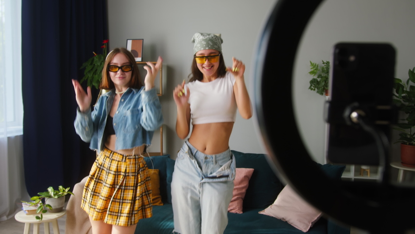 Two young sisters wearing sunglasses recording trend dance video on smartphone with lamp in living room. Female students, teenagers dancing for musical clip. Bloggers making music content for channel | Shutterstock HD Video #1076098445