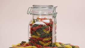 Close-up of rotating colourful macaroni in closed glass jar on white background. Shooting of color pasta in studio. Macro footage of pasta products, spaghetti. Food cooking video. 