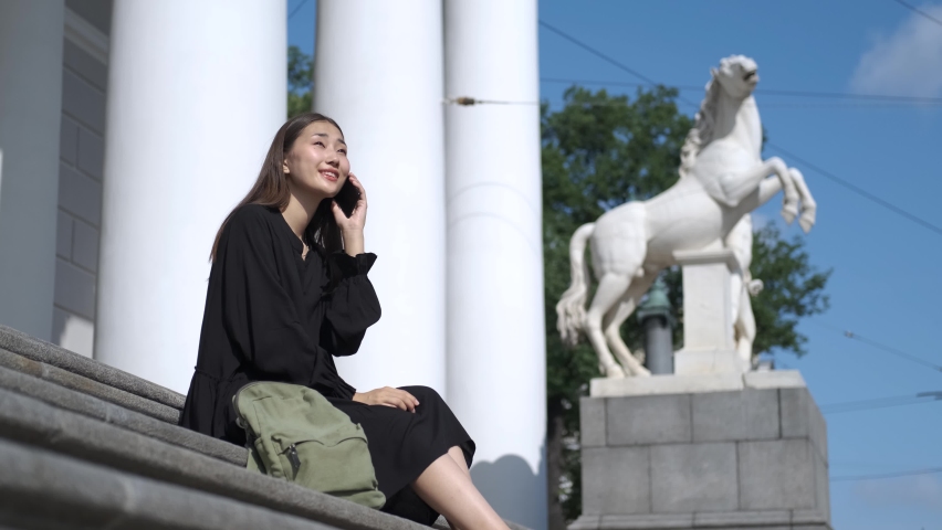 Asian woman talking on the phone while sitting on the steps against the background of the monument in St. Petersburg