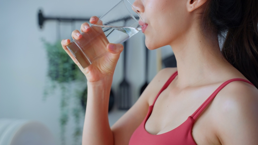 Close up of Asian attractive woman drink water after exercise in house. Young thirsty beautiful sport girl takes a sips of clean mineral natural in cup after workout for health care in kitchen at home