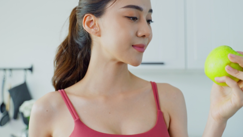 Asian beautiful woman feel happy after lose weight for health in house. Attractive girl hold green apple and smiling, look at camera while wearing big jeans in kitchen. Healthy food and Diet concept.