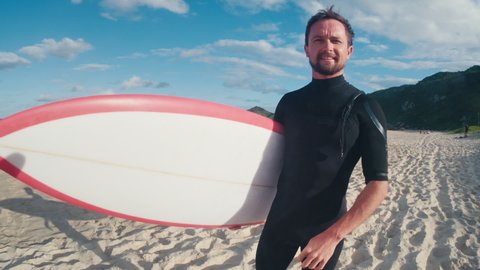 Surfer with board. Male caucasian surfer in black wetsuit stands on the beach and holds the surf board