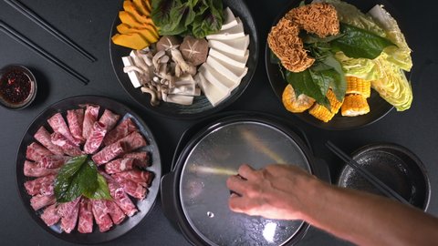 Сhef's hand opening the lid of a pot of steam. Chinese hotpot with pieces of fresh meat, instant noodles, corn, mushrooms and vegetables on a black background. Top down view. Asian food concept. : stockvideo