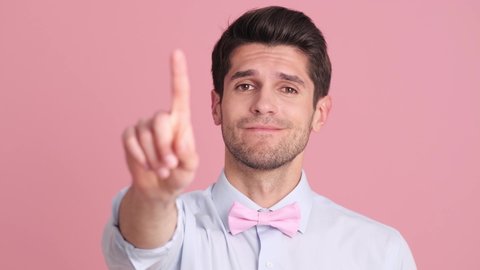 A serious young man wearing a bow tie is shaking his finger doing "not so fast" gesture standing isolated over a pink wall in the studio