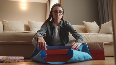 The smiling woman in glasses wrapping a gift in gift paper while sitting on the floor in the room