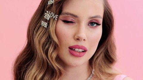 A close-up view of a pleased young woman wearing gorgeous jewelries and hairpins is winking to the camera standing isolated over a pink wall in the studio