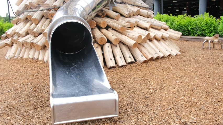 Trendy metal slide on an eco wooden playground. Empty public space with no one, slide on designed metal playground with brown wooden decorations against green bushes