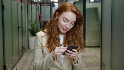 Close-up smiling attractive businesswoman using smartphone . Portrait of confident woman executive with red hair in formal wear using app on mobile phone while standing in office corridor.