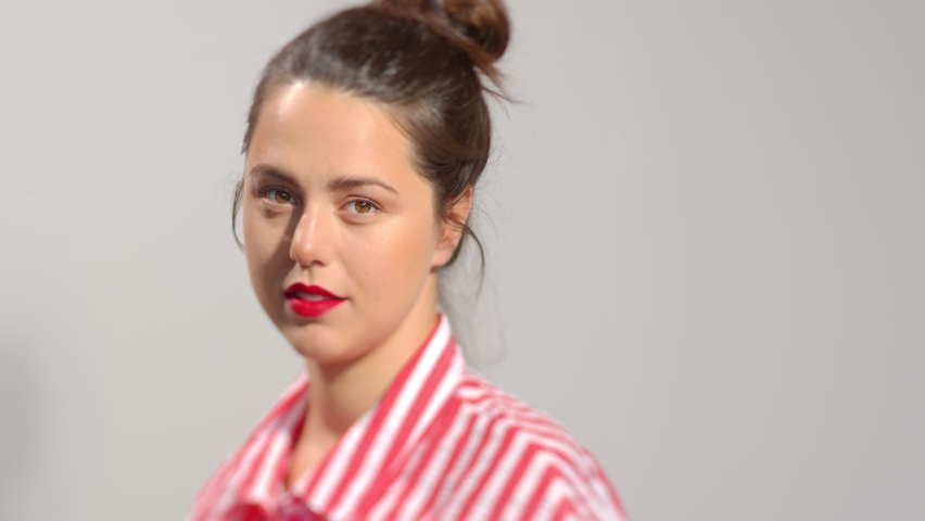 Young attractive brunette with red lipstick close up video portrait. Dark hair girl smiles lookin to the camera wide smile showing teeth. Studio still shot slow motion.