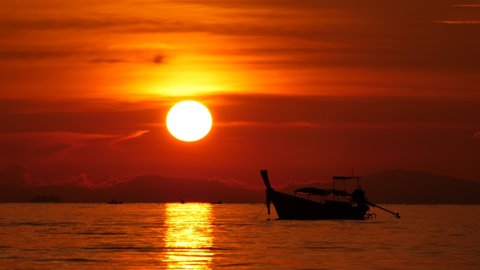 Iconic view of topical sunset in Thailand, large sun disc hang low, boat silhouette on foreground. Few tourists kayaking at calm water of sea bay at distance
