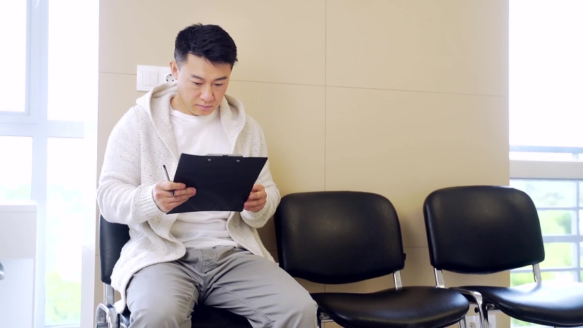 young asian man student sits on a chair in the waiting room, fills out application form. Male in hospital reception or office at employment or before treatment or testing. Patient Expected in hallway Royalty-Free Stock Footage #1076142308