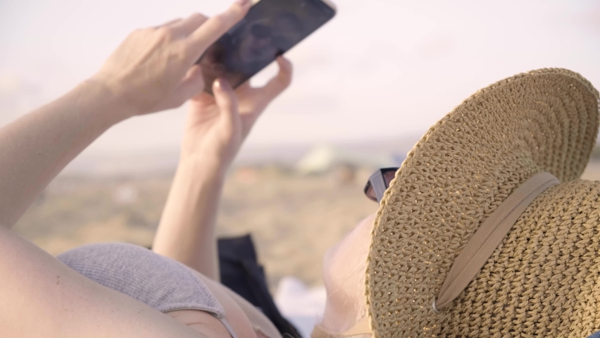 Woman taking a selfie on the beach using a smartphone laying on the sand. Girl shooting a video for social media. smartphone. posing to the camera summertime afternoon. Vlogger taking a reel or story