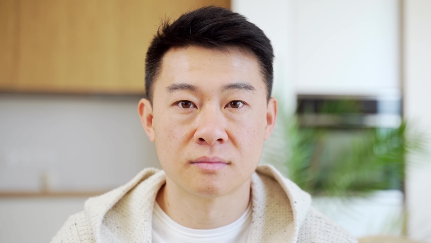 Close-up headshot portrait, face of Asian man looking at camera indoors. Face The young male in the room looks seriously and confidently. People. In casual clothes at home. Closeup head shot Royalty-Free Stock Footage #1076156147