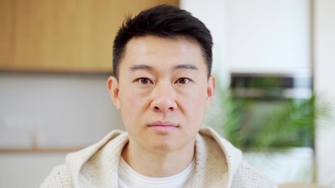 Close-up headshot portrait, face of Asian man looking at camera indoors. Face The young male in the room looks seriously and confidently. People. In casual clothes at home. Closeup head shot