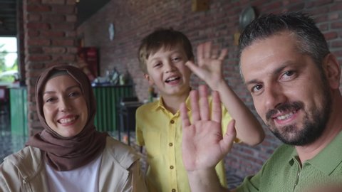 Bearded young man with his stylish wife and little son in hijab is shooting video with his cell phone camera. Young family laughing and waving at the camera.Frontal phone camera view.