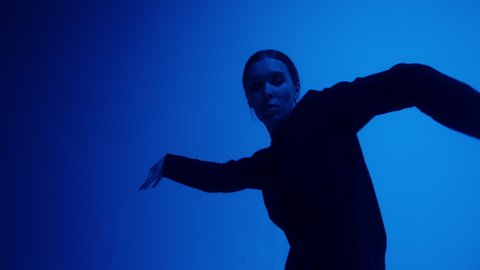 Attractive talented woman dancing hip hop on blue neon background in studio. Shooting of modern choreography for advertising of dance school, dancer expressing feelings for music video performance.