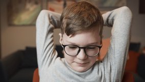 Portrait of small tired boy wearing circle glasses doing warm-up exercises, stretching his hands and back. Child rubbing his eyes after long study at computer during the coronavirus isolation at home.