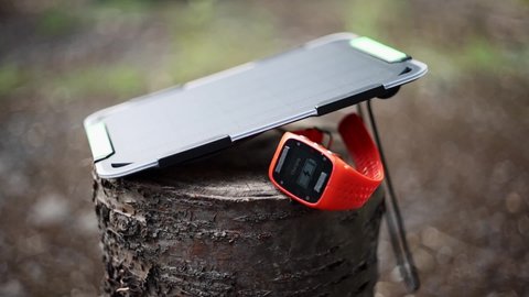 Concept of renewable energy. Portable solar panel charges a smartwatch on a tree stump in a summer day. 