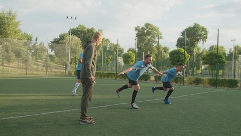 Soccer players in football sportswear standing on bosu ball, balancing on feet under control of young sport instructor outdoors, while player of multiracial team kicking ball on background at sunrise