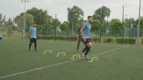 Football young instructor controlling performance of jump barriers exercise by soccer team of mixed race sportsmen in sportswear outdoors at daybreak. Soccer training of players on football pitch - Powered by Shutterstock - Get 15% off with code: PIKWIZARD15