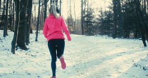Running woman, headphone girl runner on the snow in winter sunny day. Female in pink sportswear jacket fitness training outdoors. Sport concept, leisure and freedom. 4K slow motion video 