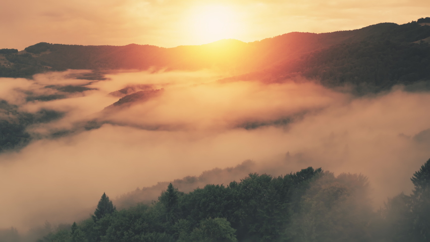 Aerial view, dense fog on mountain tops, bright orange sunrise. Yellow sunset over gray haze and evergreen forest on hills. Magical summer forest, sun rays. Nature concept. Cinematic drone flight