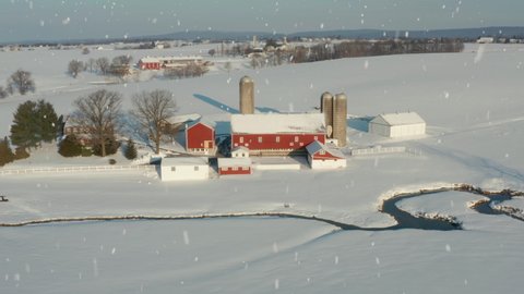 Winter snow with red barn farm buildings. Aerial of white snowflakes falling on field and stream.
