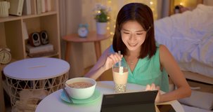 asian woman is eating instant noodles and tapioca ball milk tea while watching video on digital tablet at home in the evening