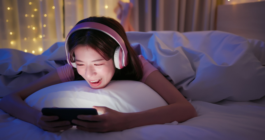 Asian woman wearing headsets play mobile games and win with fist gesture by smartphone on bed at night | Shutterstock HD Video #1076179157