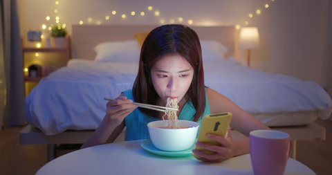 asian woman is eating instant noodles while looking at social media on mobile phone in the evening