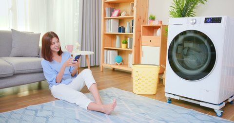 asian woman lying on floor do the laundry in smart washing machine and use smartphone operate it at home