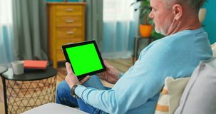 Adult Old Man at Home Uses Green Mock-up Screen Tablet. He is Sitting On a Couch in His Cozy Living Room. Over the Shoulder Camera Shot