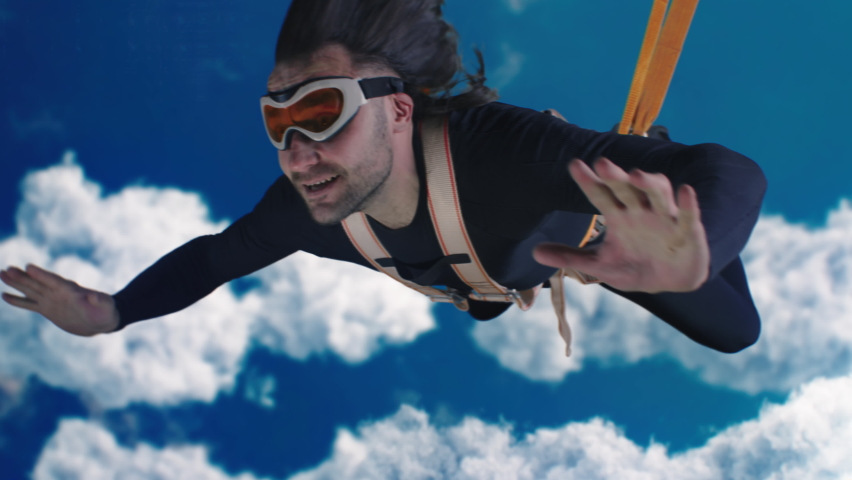 Adult man in protective gear screaming in excitement and gesturing thumbs up while falling down with parachute in blue cloudy sky