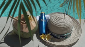 Video footage of green young coconut close up with bamboo straw, bottle of sunscreen SPF 50, sun hat, tropical yellow flower frangipani, palm shade on the edge of swimming pool in Bali