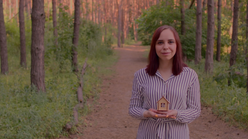 Young woman with small wooden house in hands stands in forest. Concept of purchasing new apartment and roof overhead.