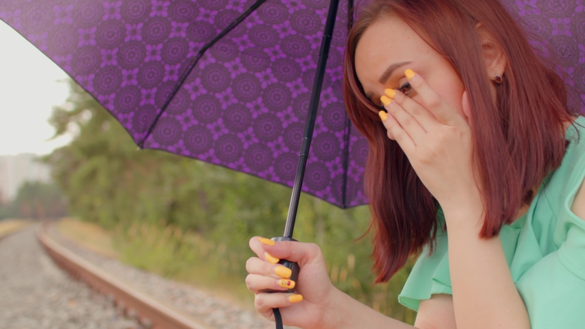 Young woman with umbrella rubs her eyes, sitting on railway in cloudy and windy weather. Adult female scratches eyes, sitting on rail in countryside.
