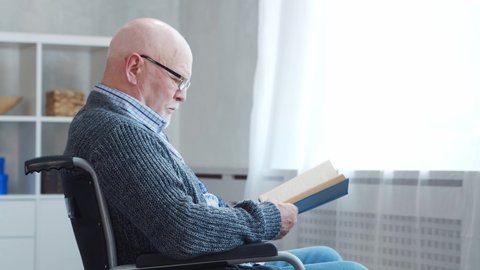 Disabled old man is sitting in a wheelchair at home alone. A handicapped person in a wheelchair is reading the book. Paralysis and disability.