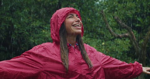 Authentic shot of carefree young woman wearing red protection cape is feeling free and smiling under the rain on a background of green trees. Concept of life, freedom, nature, adventure, purity.