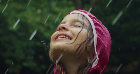 Authentic shot of carefree happy little girl in raincoat is having fun and enjoying the rain in a hot summer day in a green park. Concept: freedom, childhood, happiness, love for, nature, life