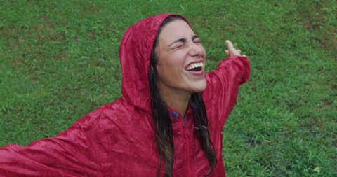 Authentic shot of carefree young woman wearing red protection cape is feeling free and smiling under the rain on a background of green trees. Concept of life, freedom, nature, adventure, purity.