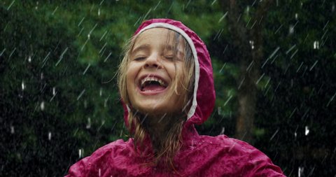 Authentic shot of carefree happy little girl in raincoat is having fun and enjoying the rain in a hot summer day in a green park. Concept: freedom, childhood, happiness, love for nature, life