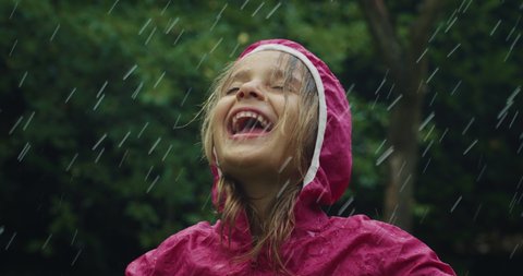 Authentic shot of carefree happy little girl in raincoat is having fun and enjoying the rain in a hot summer day in a green park. Concept: freedom, childhood, happiness, love for, nature, life