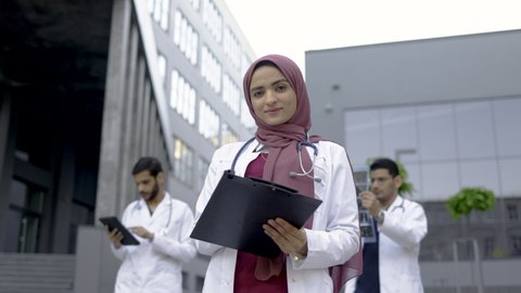 Female Muslim pleasant doctor in hijab, standing with clipboard outdoors in front of hospital building, while two handsome Arabian men doctors working with x-ray and tablet pc behind