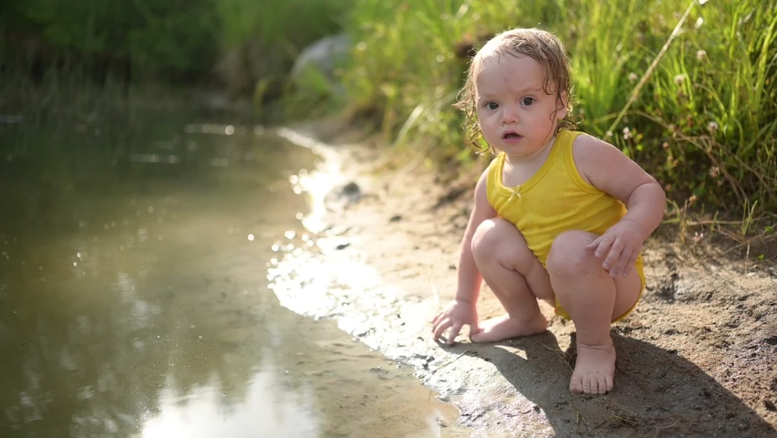 Little funny cute blonde girl child toddler in yellow wet bodysuit playing by the lake waterside shore at sunset outside. Baby with mud on hands and legs. Water activities at summer