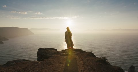 Fairy tale little girl with long hair goes to the edge of cliff to look stunning seascape at early morning. Girl character meeting sunrise on high mountain near sea. Wonderland landscape view Adlı Stok Video